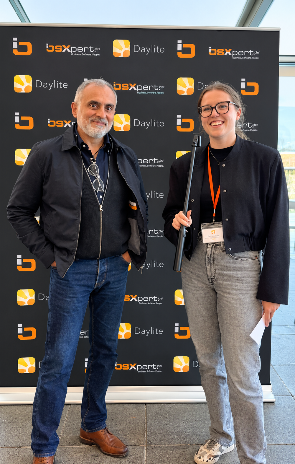 Eine Frau interviewt einen Mann. Beide stehen vor einer schwarzen Leinwand auf der orangene Daylite und iosxpert Logos abgebildet sind. Beide Personen lächeln in die Kamera.